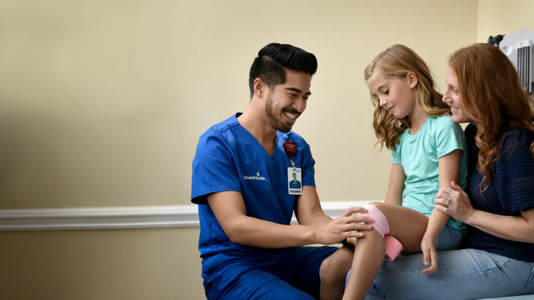 A Provider Wraps a Child Patient's Knee With Pink Gauze While a Parent Watches