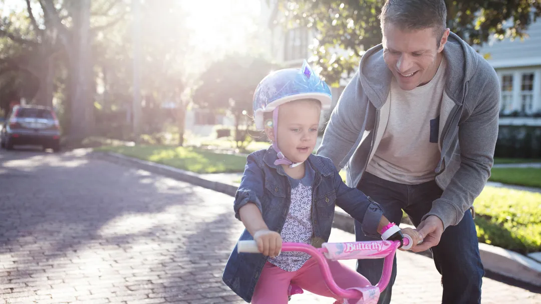 Father teaching his young daughter how to ride a pink bike in the street.