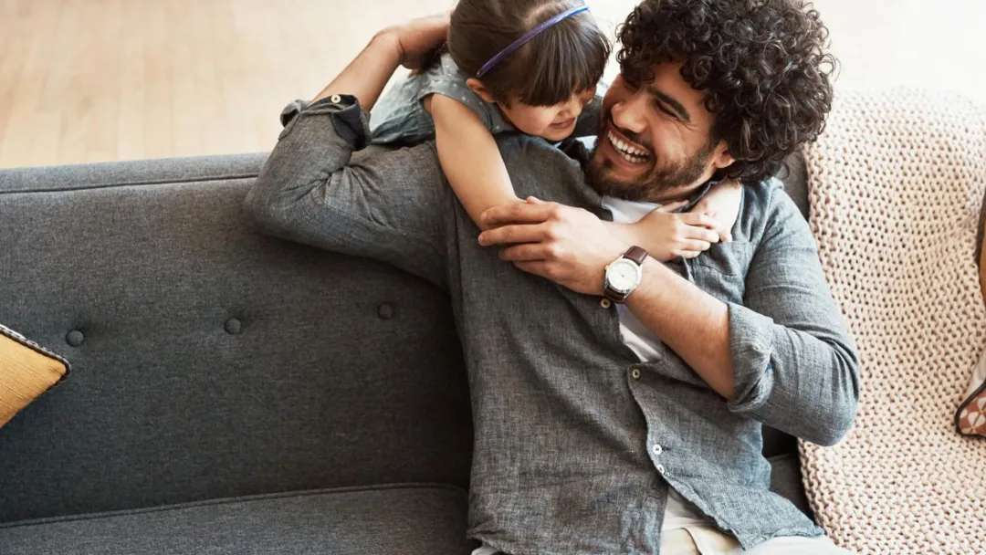 A daugther hugging her father who is sitting in the couch.