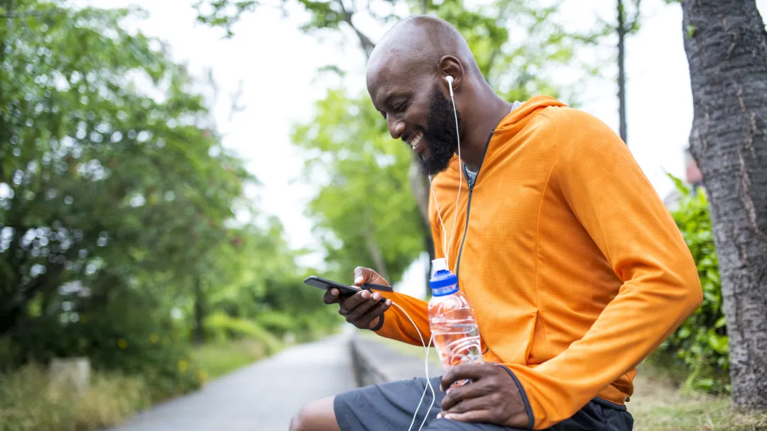 An adult man in an orange hoodie, sitting on a bench outside, looking at his phone and hydrating with a bottle of water.