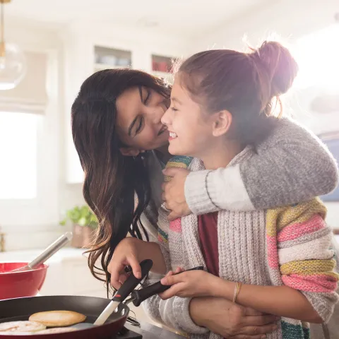A family of three women are in the kitchen, one daughter hugging the youngest while she flips pancakes.