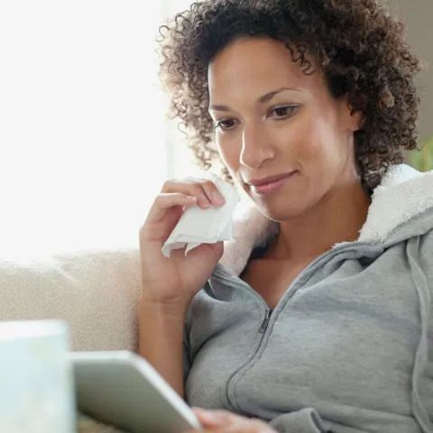 An African American woman dabs her face with a tissue while scoping out her tablet.
