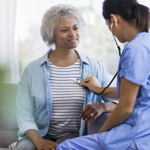 A female doctor is seated, listening to a senior woman's heart with a stethoscope.