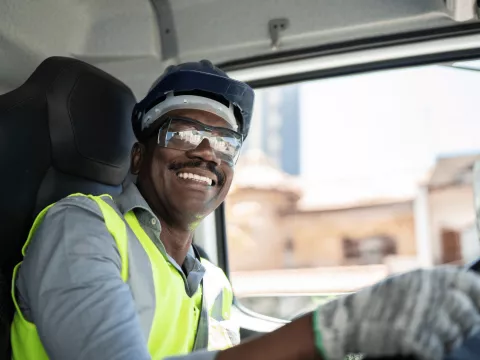 A black man wearing a safety vest, protective glasses, safety helmet and gloves while driving a truck.